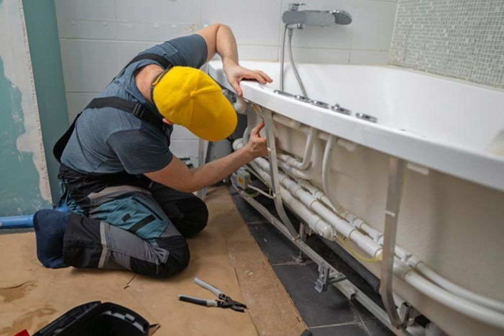 Person in yellow cap installing plumbing under a bathtub in a bathroom.