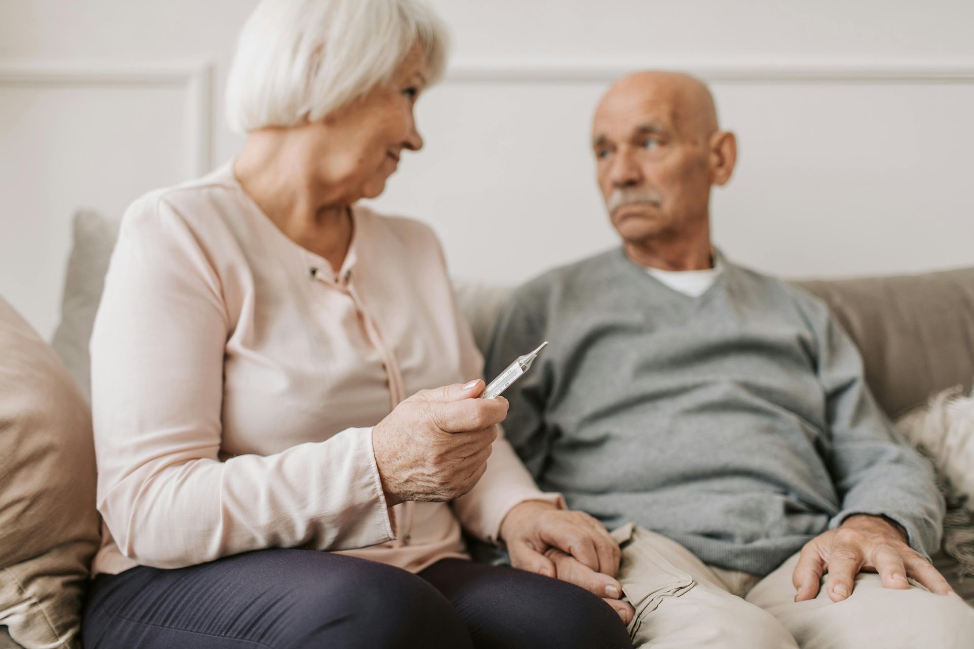 An elderly couple is sitting on a couch and the woman is holding a thermometer.