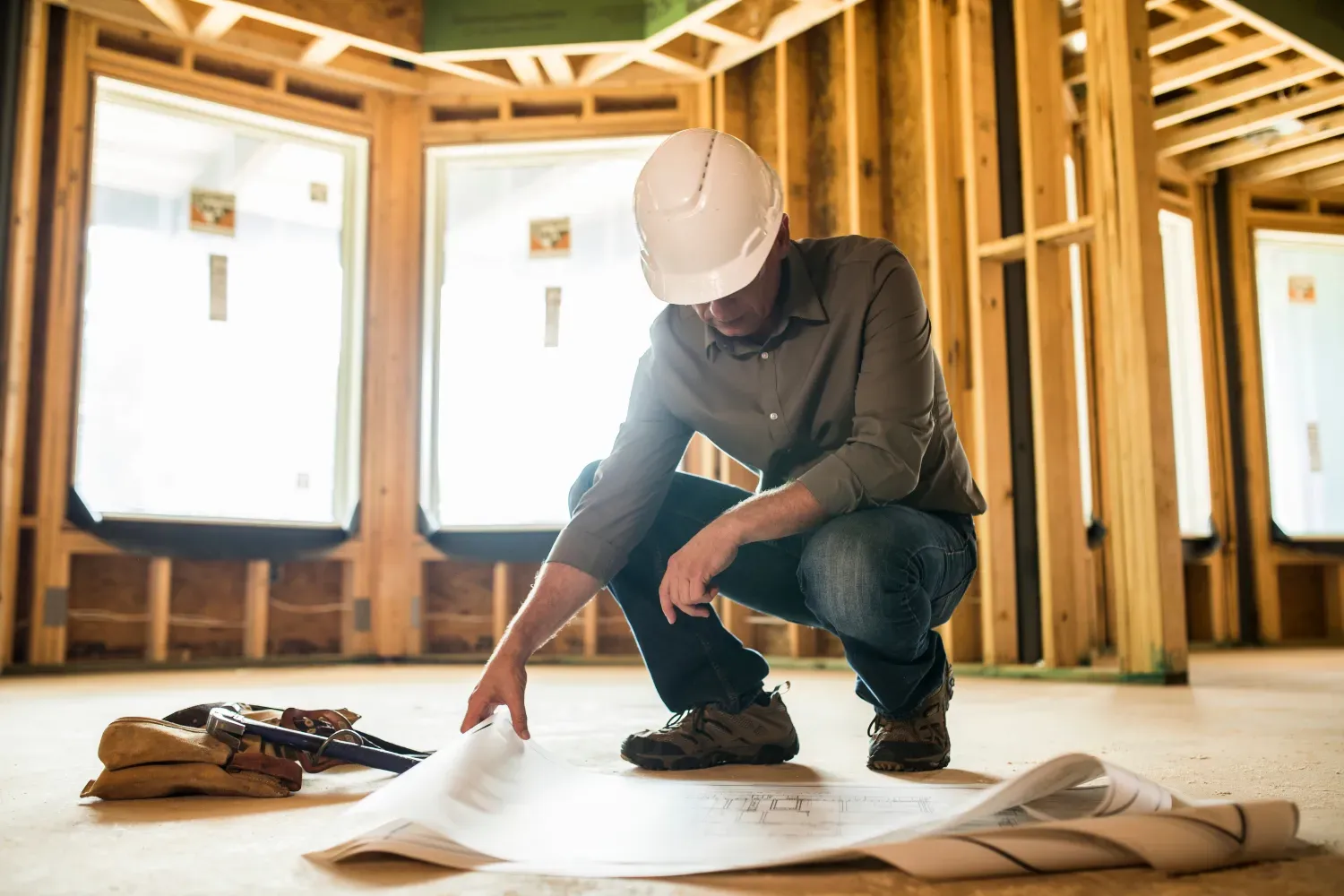A construction worker in a hard hat crouches on a wooden floor in a house under construction, reviewing blueprints.