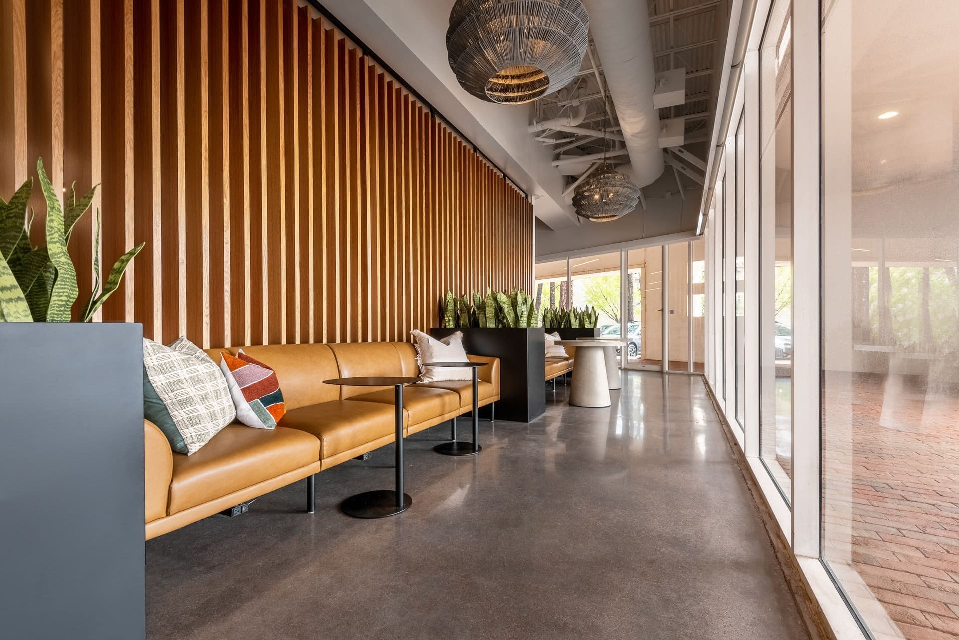 A modern office lobby with a tan leather sofa against a vertical wood slat wall, potted plants, and glossy gray floors.