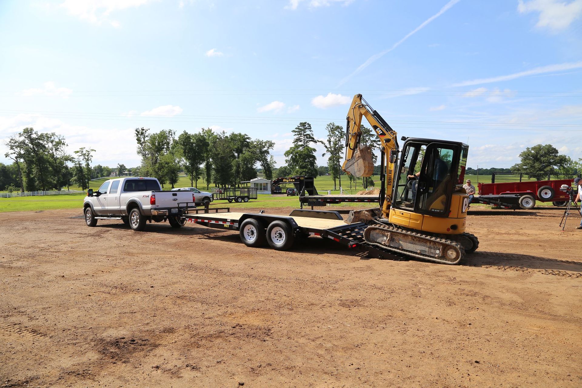 Truck towing a trailer with a small excavator on it. The scene is outdoors on a dirt surface.