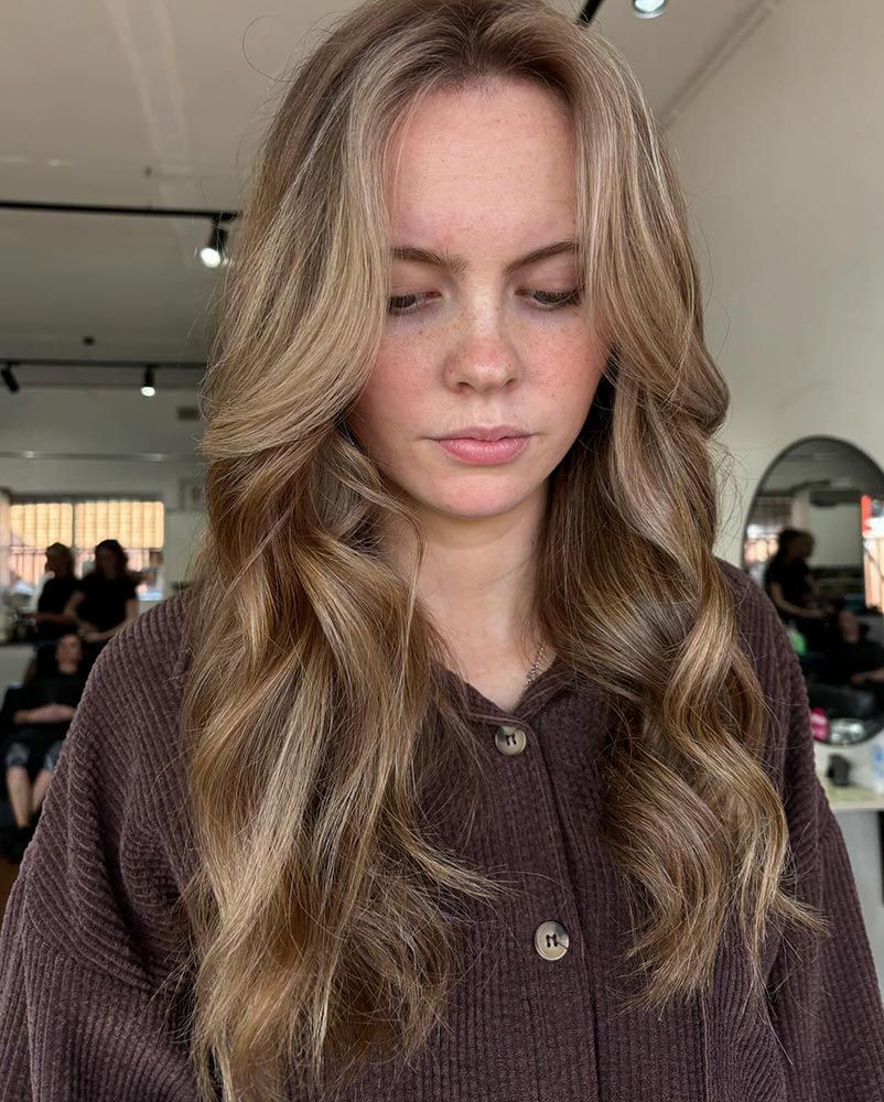 A Woman With Long Wavy Blonde Hair is Wearing a Brown Shirt — Hairenvy in Ettalong Beach, NSW