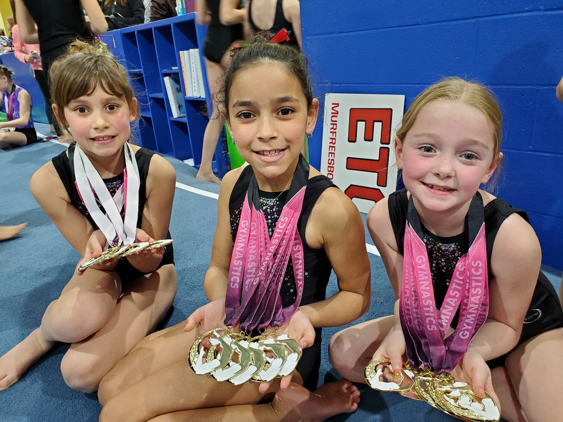 Three young girls are sitting on the floor holding medals.