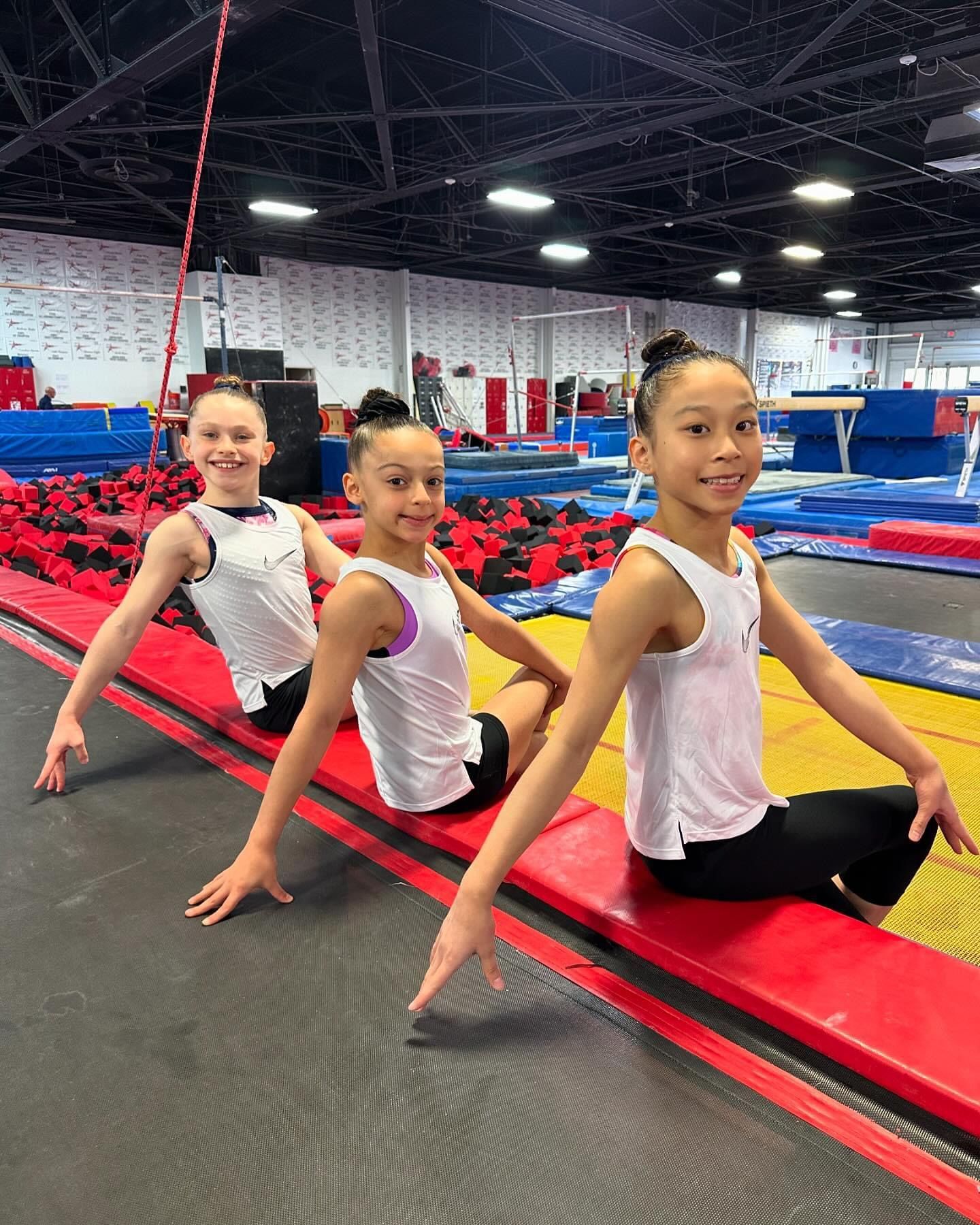 Three young girls are sitting on a balance beam in a gym.