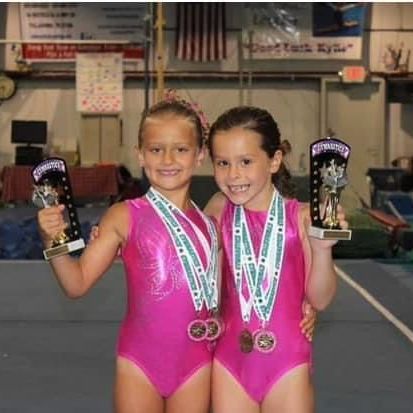 Two young girls in pink leotards are holding trophies in a gym.