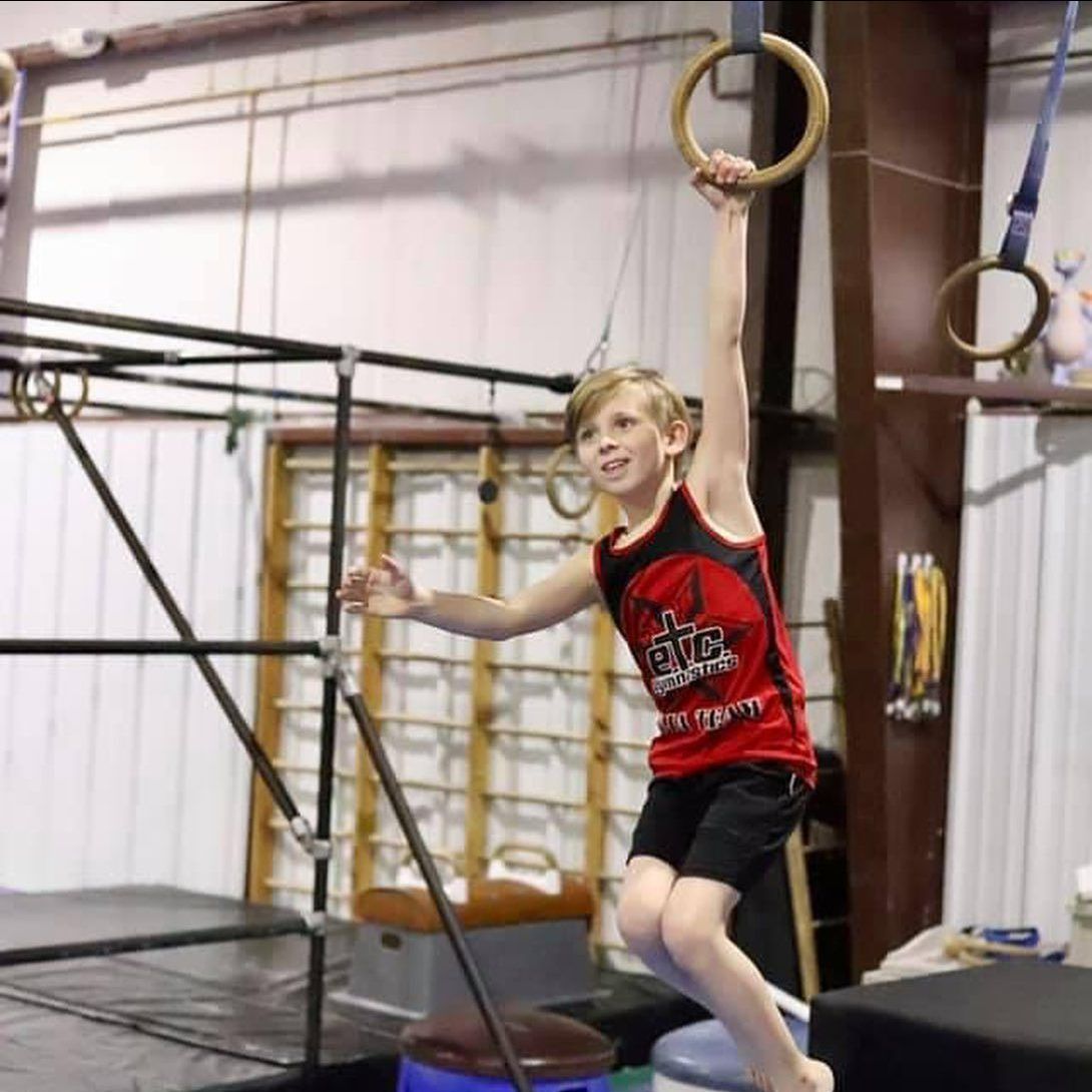 A young boy in a red tank top is hanging from gymnastic rings