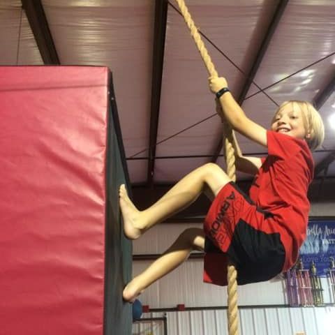 A young boy in a red shirt and black shorts is climbing a rope