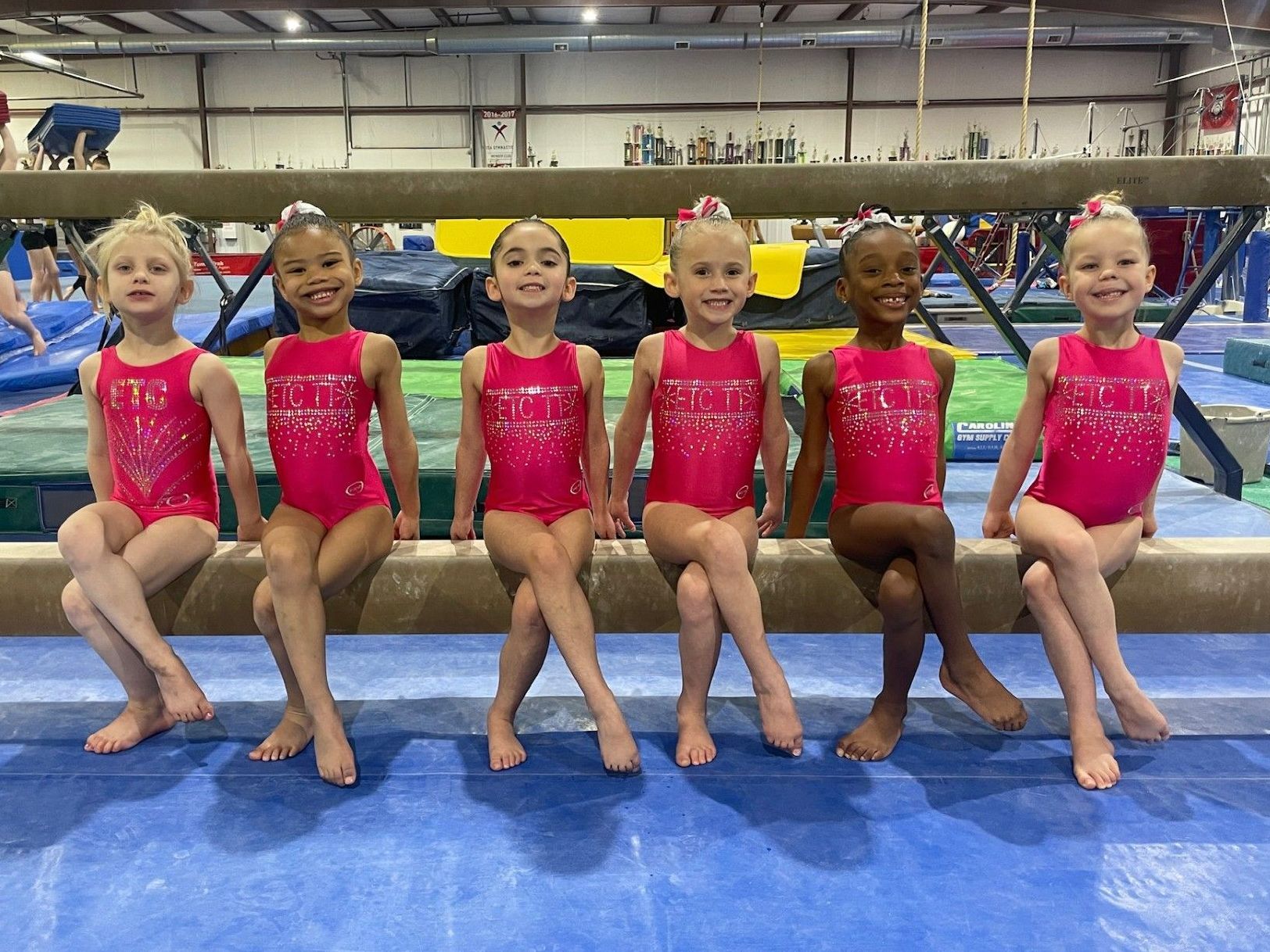 A group of young girls are sitting on a balance beam in a gym.