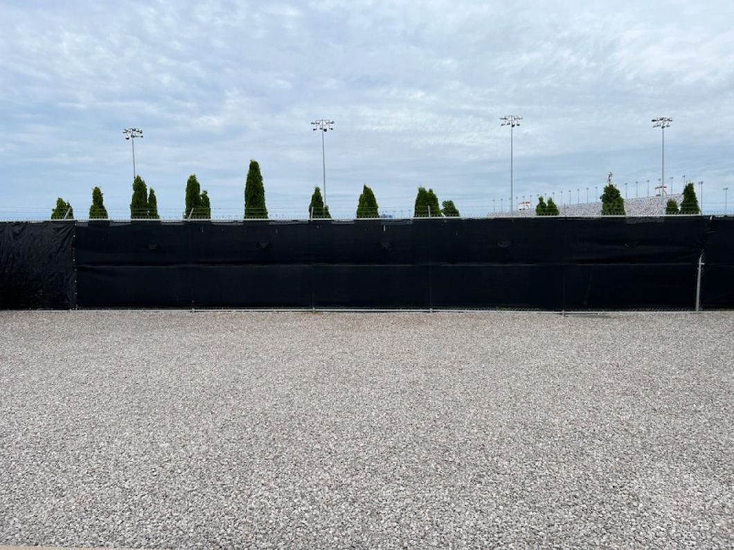 A gravel area with a black fence and trees in the background.