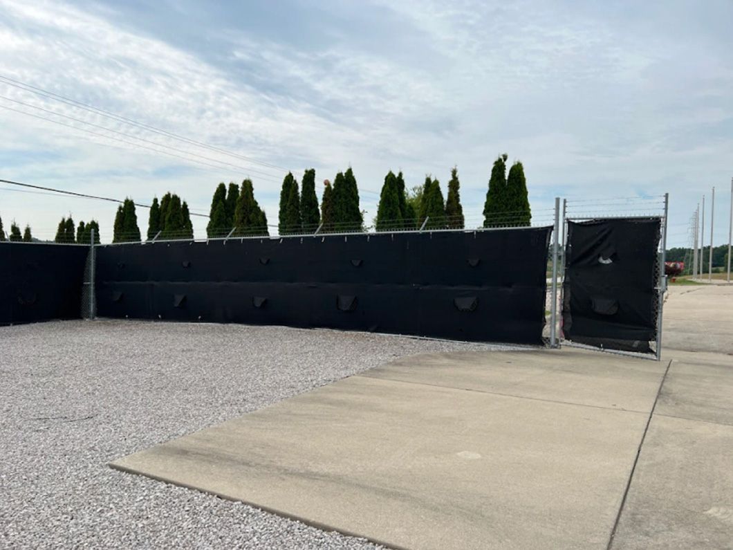 A black fence is surrounded by gravel and trees in a parking lot.