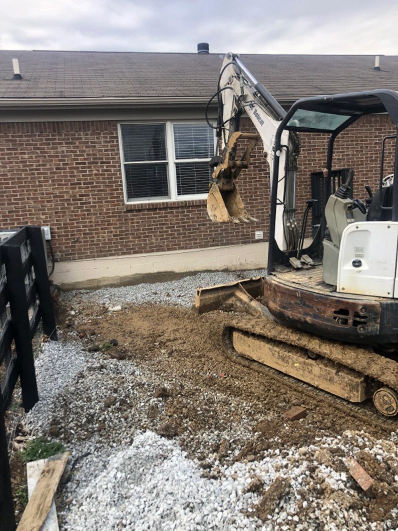 A small excavator is digging a hole in front of a brick house.