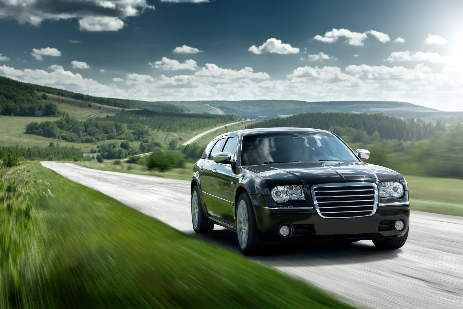 Black Chrysler sedan driving on a country road with rolling green hills under a cloudy sky.