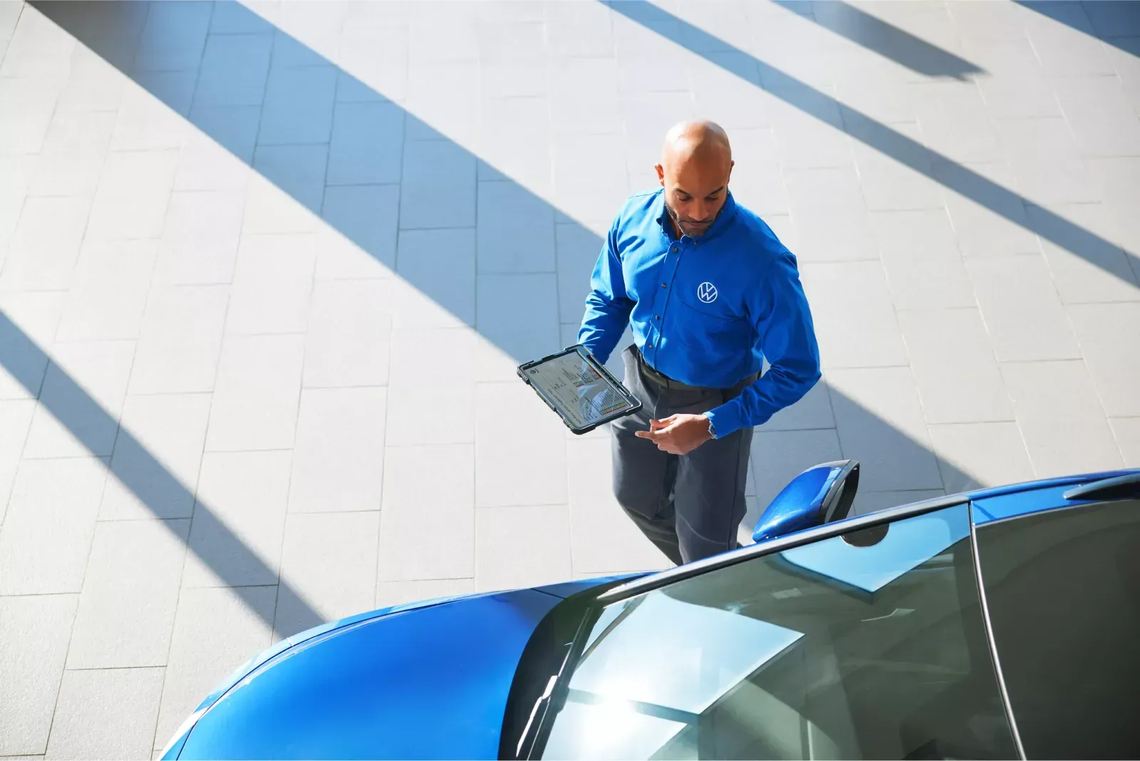 Man in blue shirt using tablet near a blue car in a bright indoor setting.