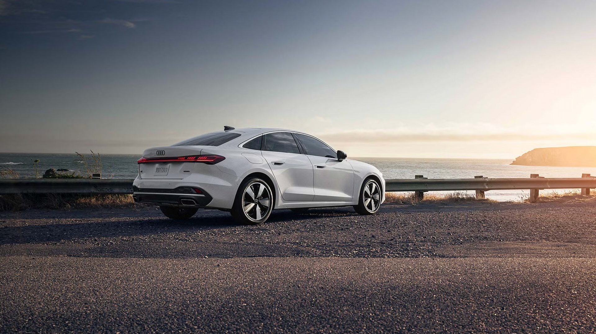 White Audi sedan parked on a coastal road, overlooking the ocean under a sunset sky.