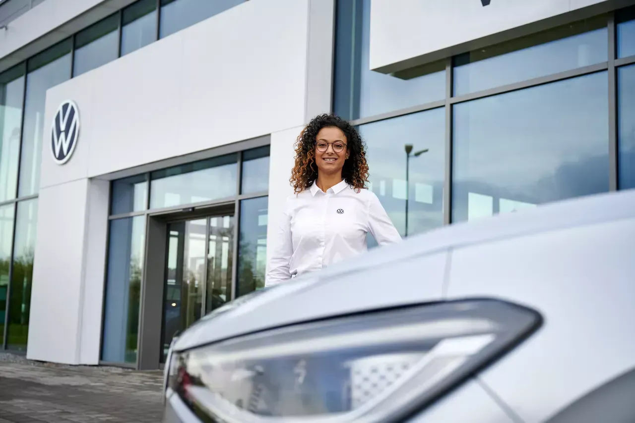 Woman in white shirt smiles next to a white car in front of a Volkswagen dealership.