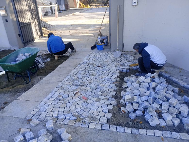 Due persone stanno stendendo un sentiero di ciottoli all'esterno di un edificio; sono presenti una carriola, degli attrezzi e dei blocchi di pietra.
