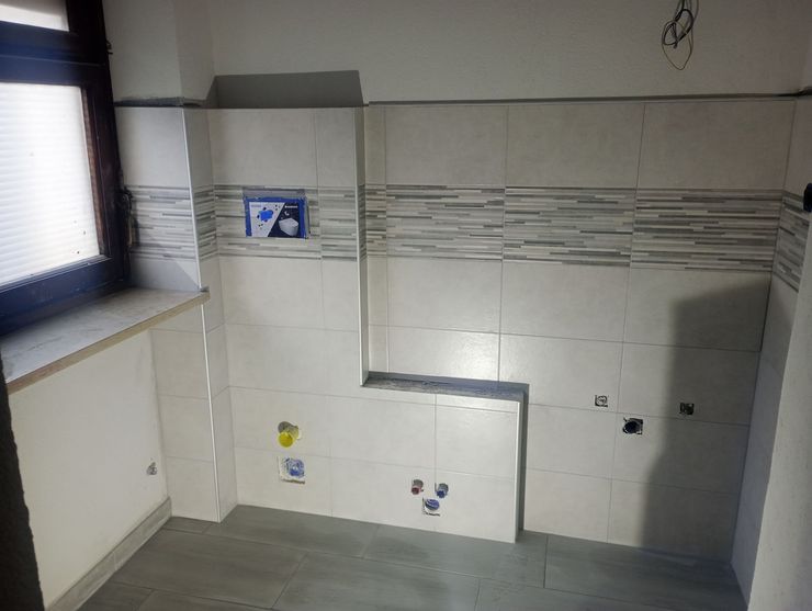 Bagno in costruzione; pareti piastrellate bianche, pavimento grigio, finestra, impianti idraulici parzialmente installati.