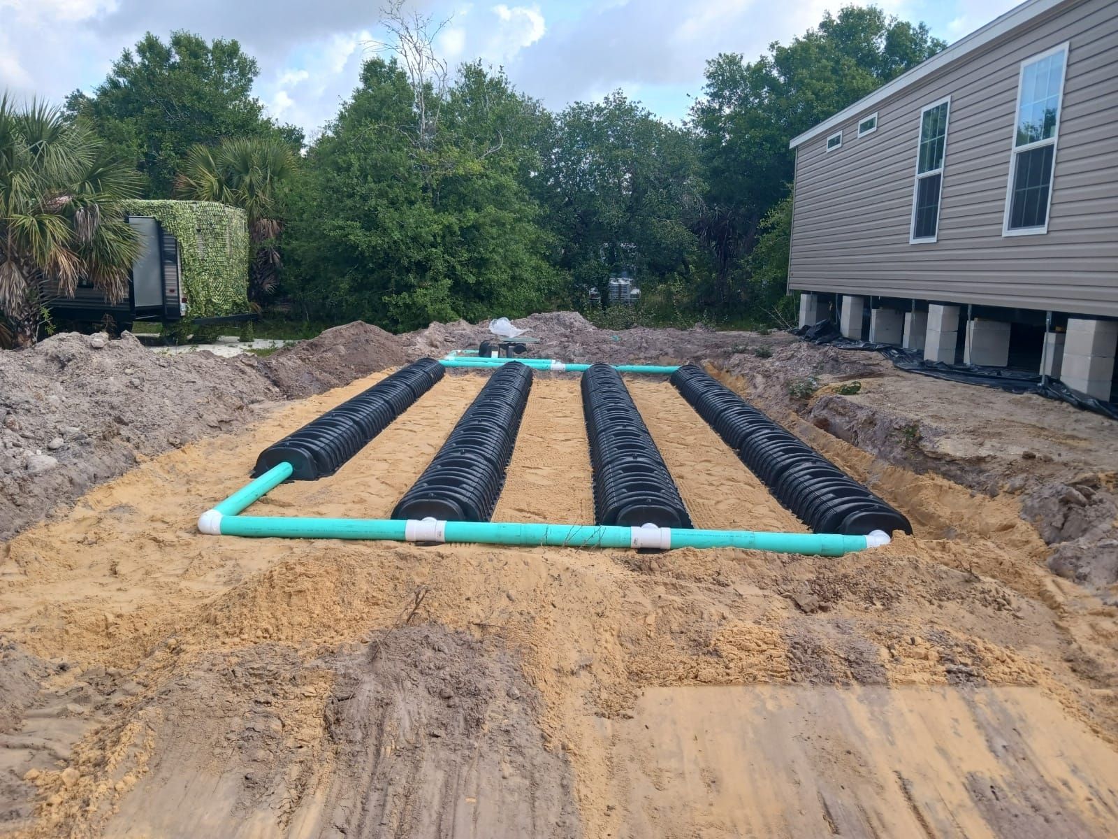 Septic system installation: black drainfield tubes laid in sand, connected by green PVC pipes, near a house.