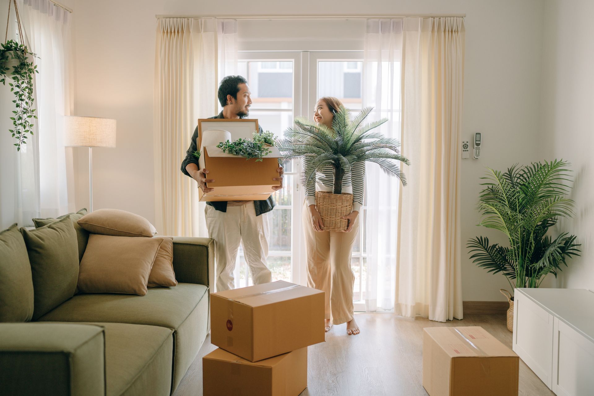 Couple carrying boxes and plants, moving into a bright, new home with a sofa and windows.