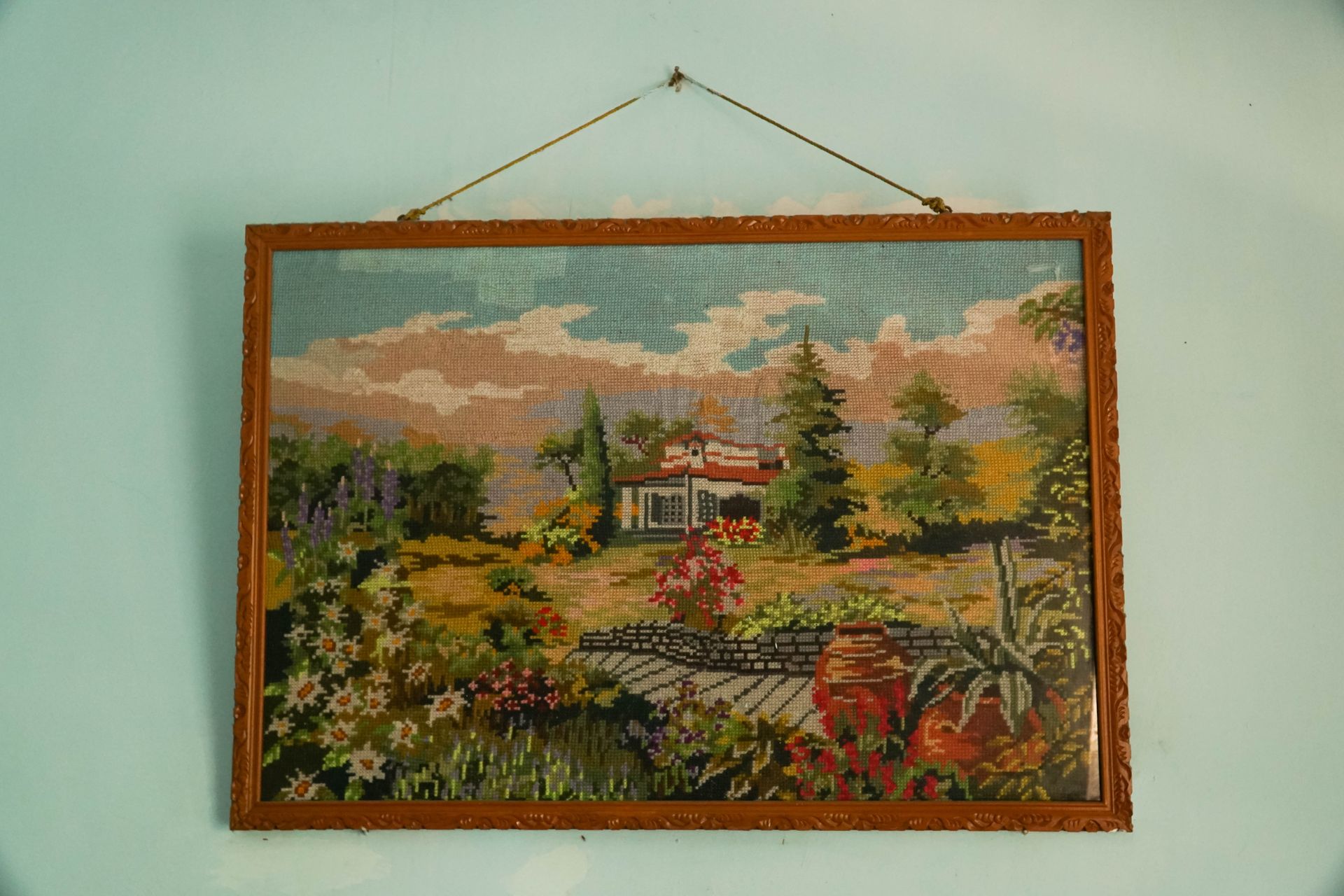 Framed needlepoint landscape: house in a garden with flowers, trees, and a blue sky.