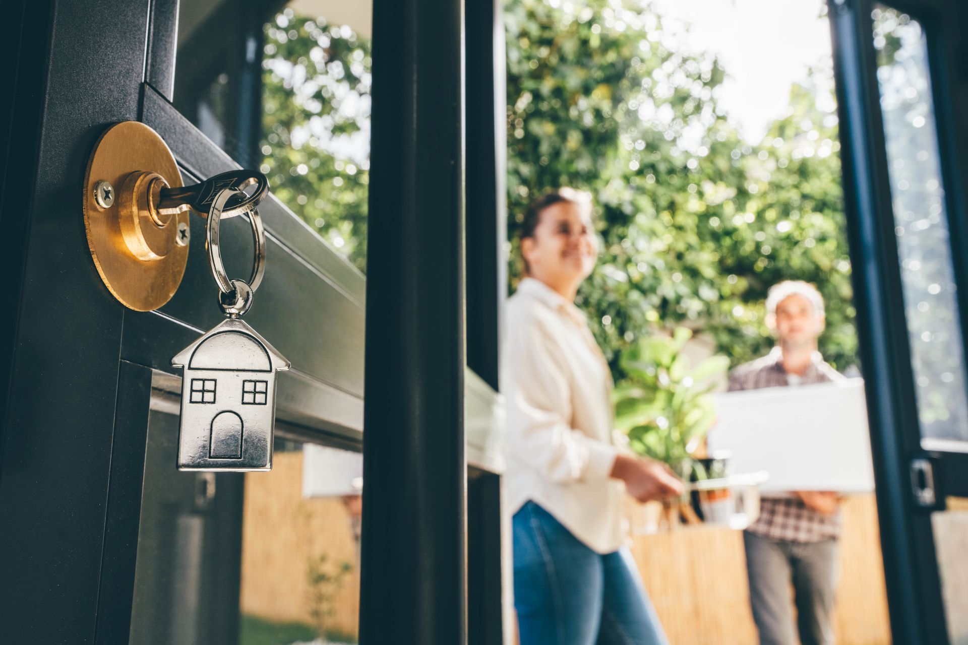 Key in door, house-shaped key chain, couple moving in with boxes and plants.