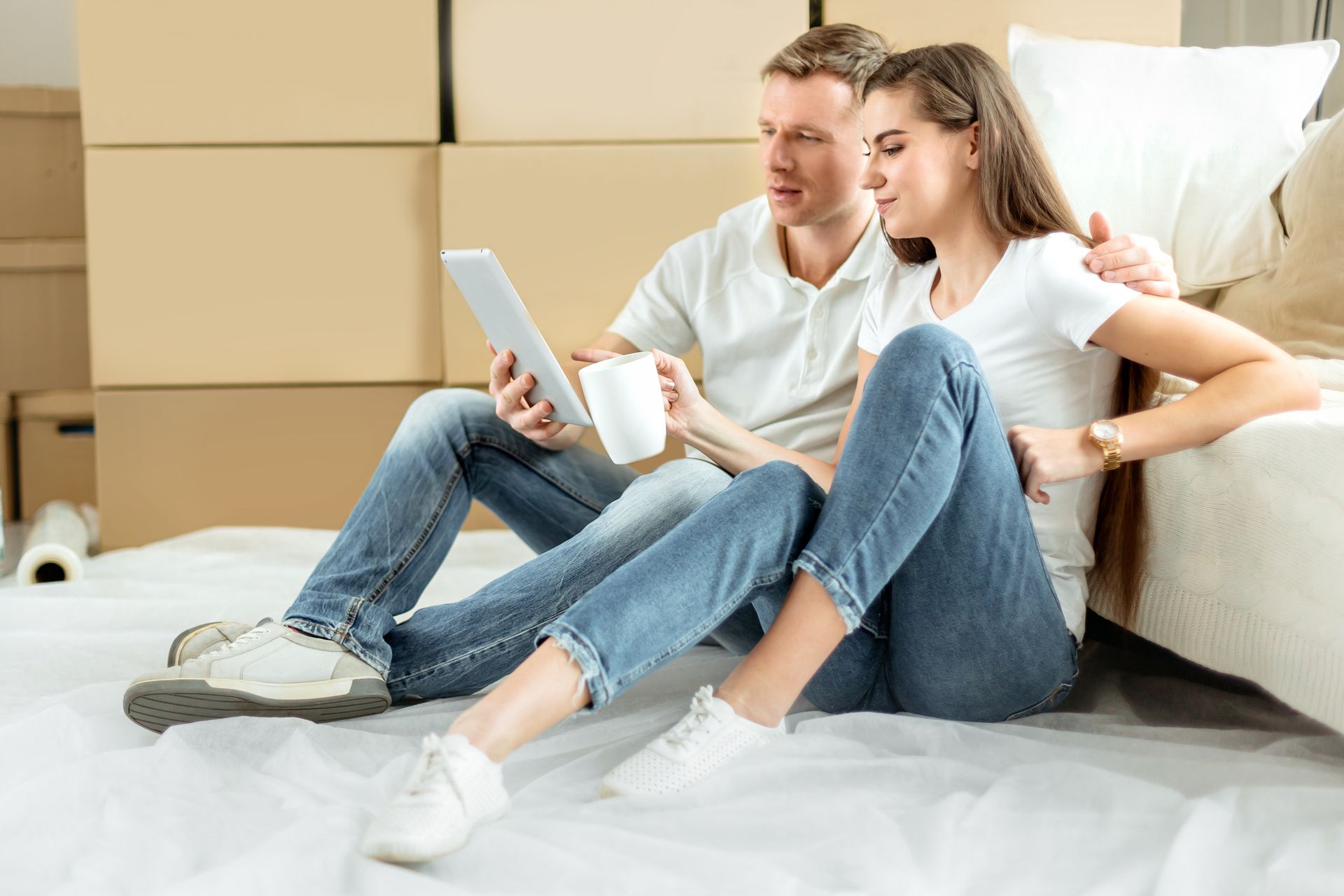 Couple sitting on floor with tablet, surrounded by moving boxes, discussing plans.