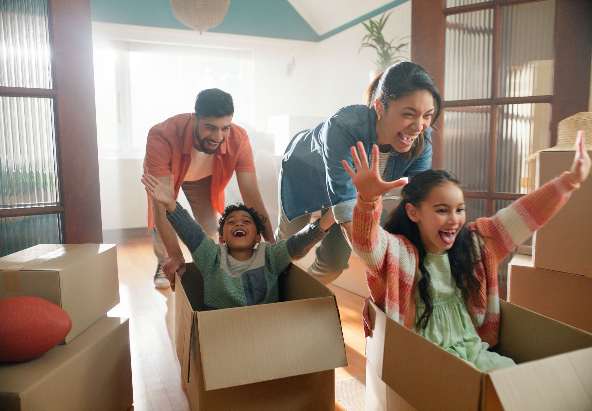 Family of four, parents pushing children in cardboard boxes, laughing in a new home.