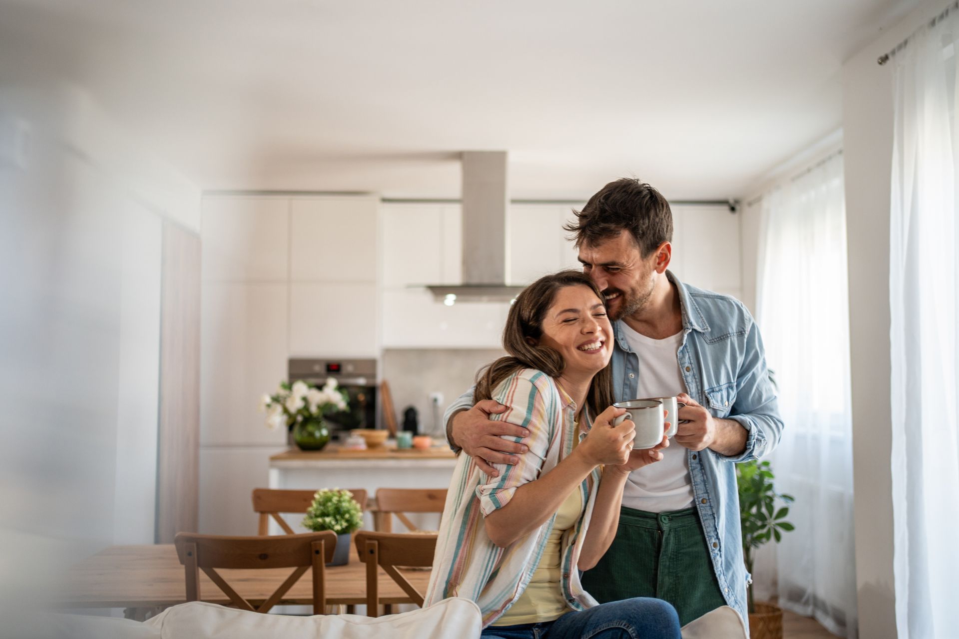 Couple in modern kitchen: man hugs woman drinking coffee, smiling; bright interior.