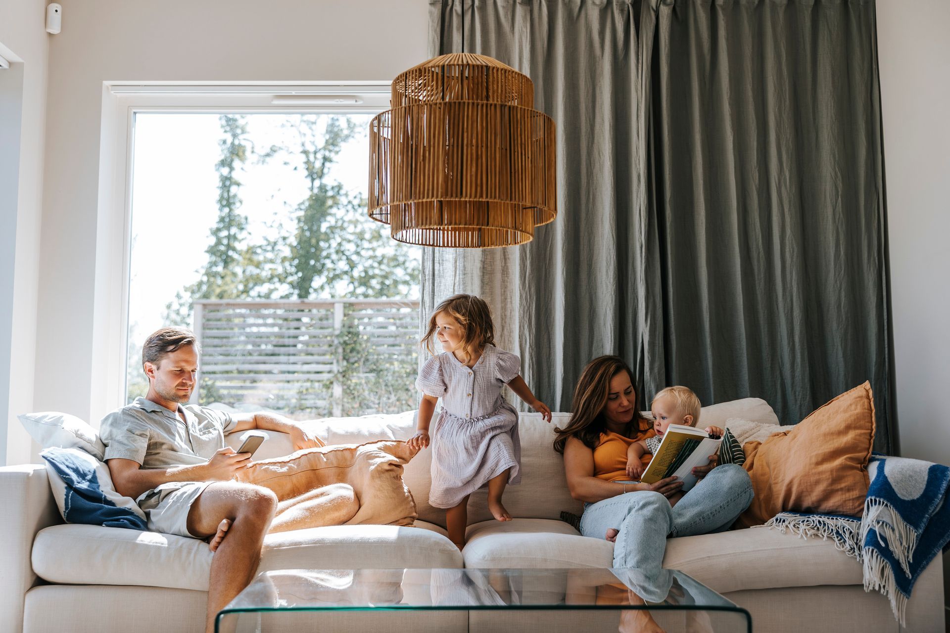 Family relaxing on a white couch in a sunny living room. Woman reading, man on phone, children playing.