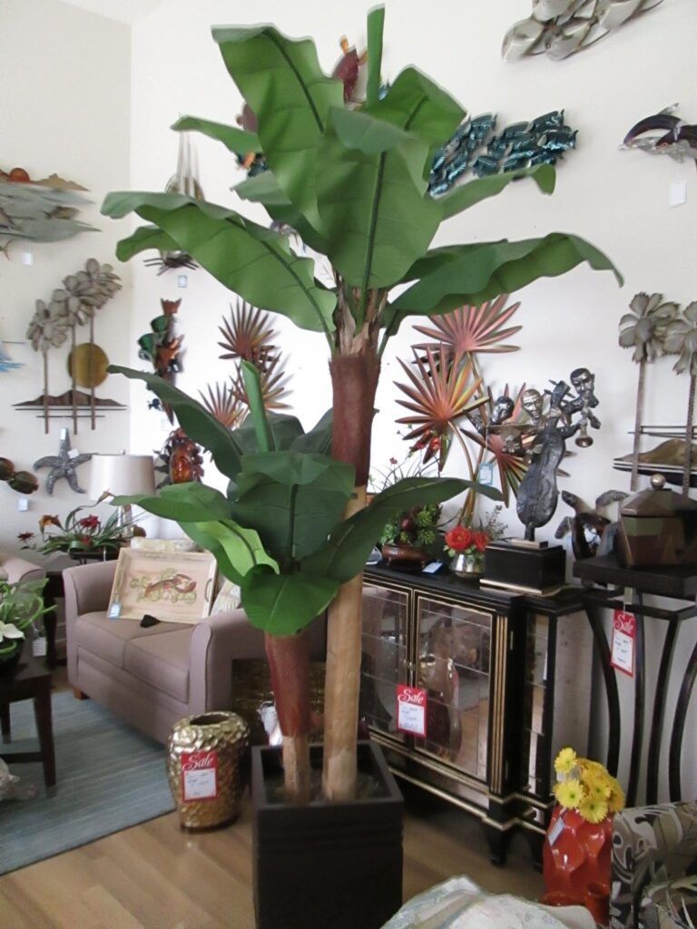 Artificial banana tree in a black square planter, surrounded by furniture and decor in a store.