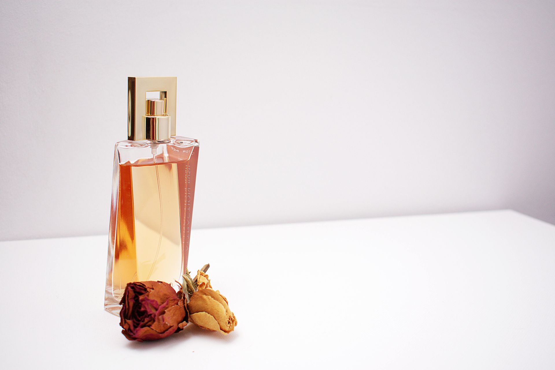 A glass perfume bottle from Scent Room stands on a white surface next to two small, dried roses.