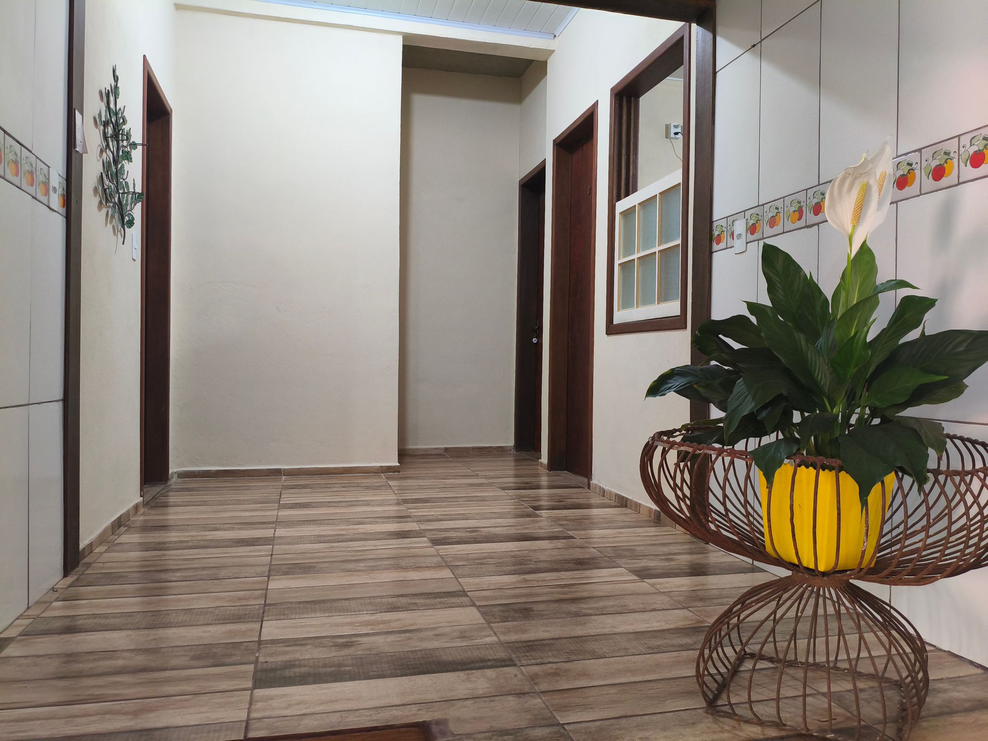 Corredor com piso de azulejo claro, paredes bege e portas marrons. Uma planta em vaso está em um suporte de metal decorativo.