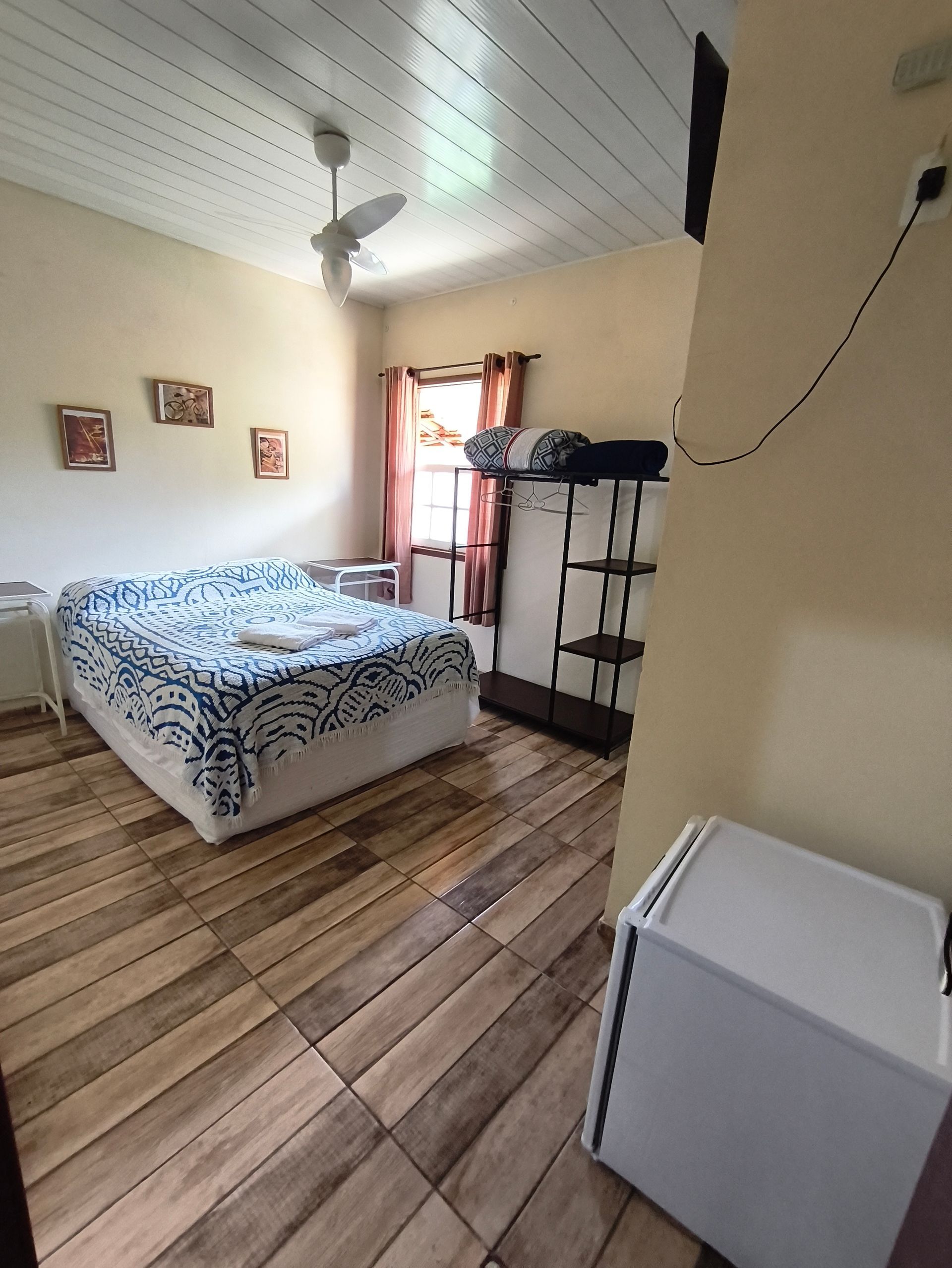 Quarto com cama, prateleiras, frigobar, janela, ventilador e piso de cerâmica imitando madeira.