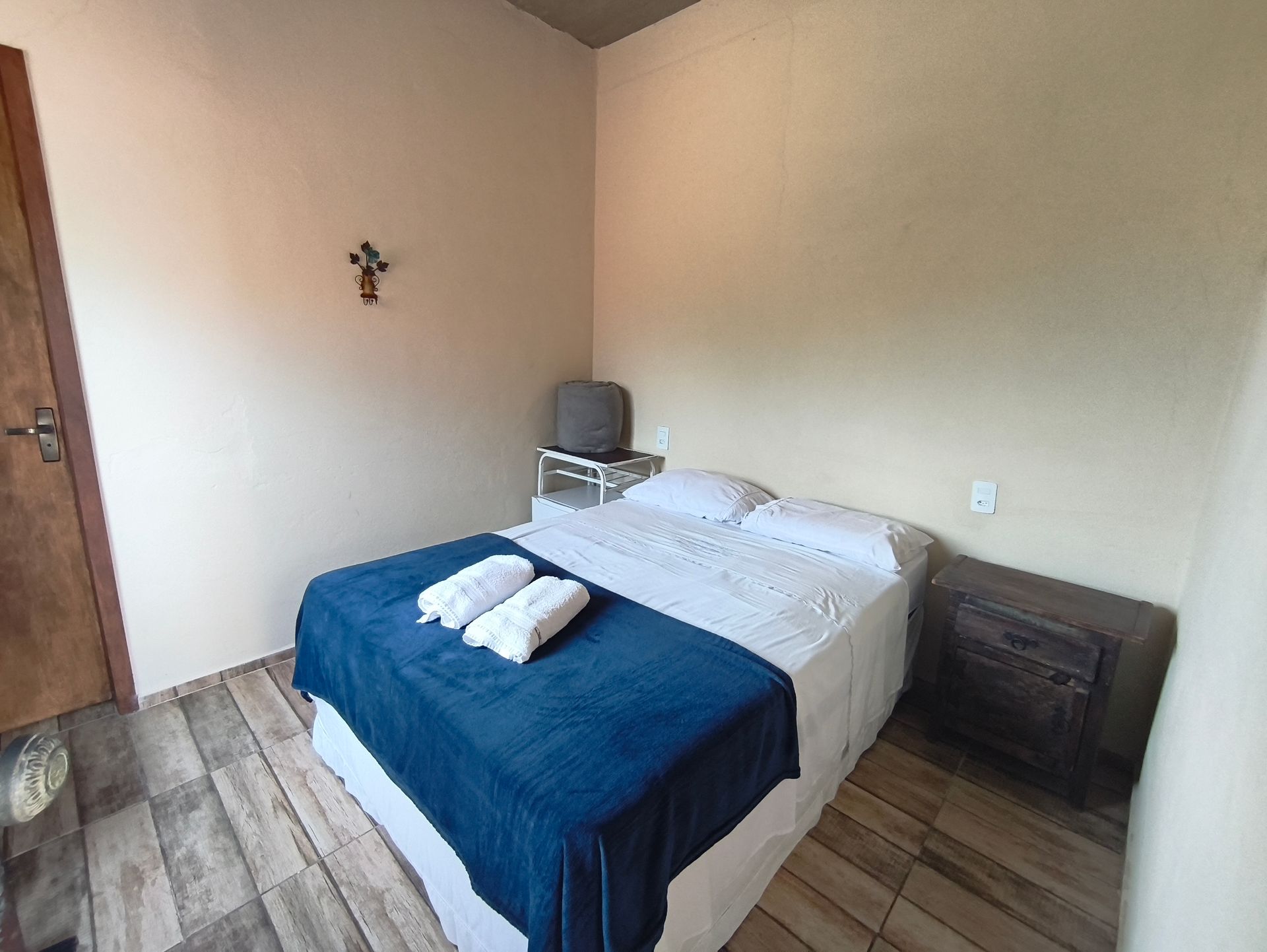 Quarto com cama, mesa de cabeceira e TV pequena. Cobertor azul e toalhas brancas sobre a cama. Paredes claras e piso de madeira.