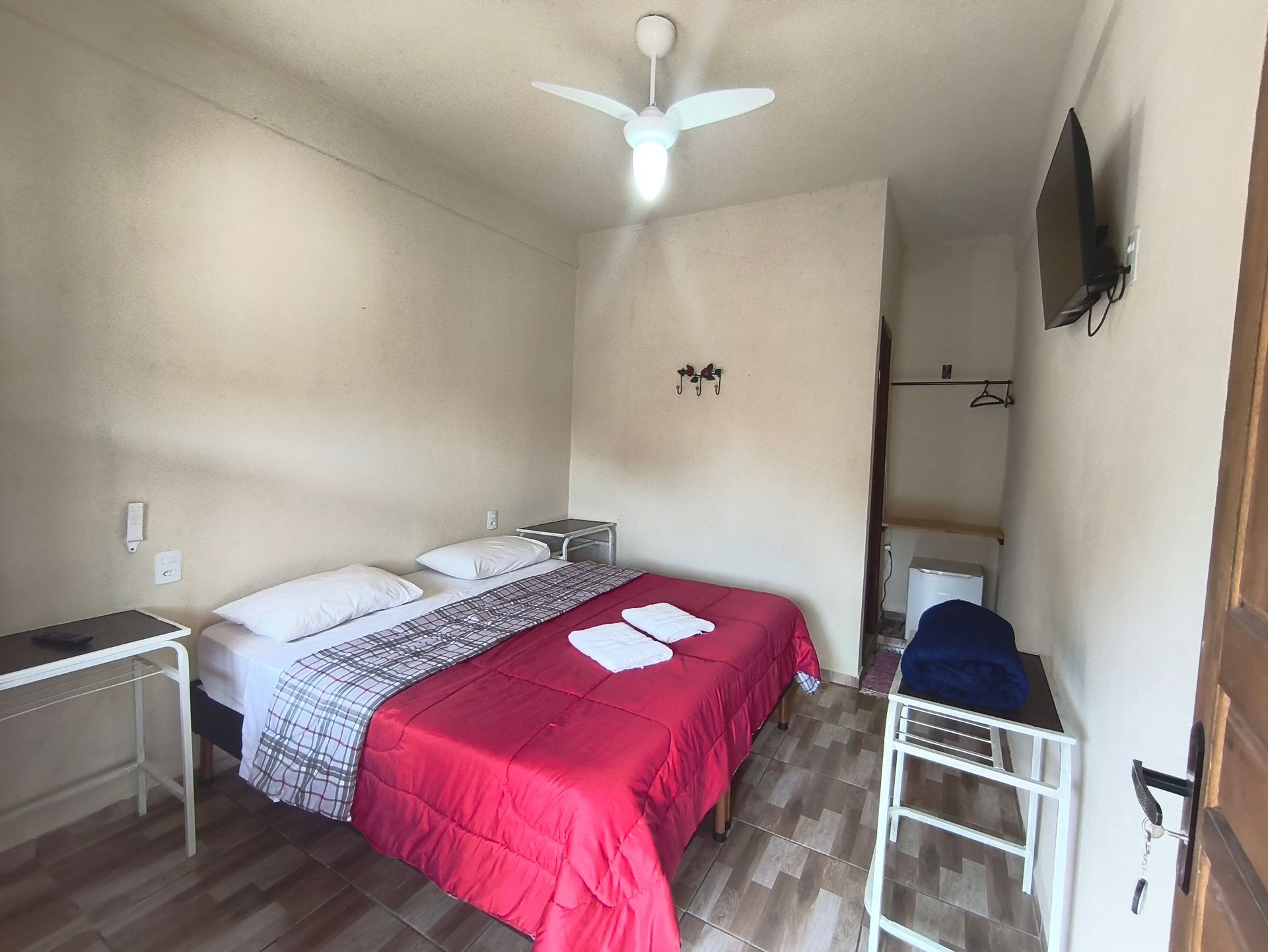 Quarto com colcha vermelha, mesas de cabeceira, TV pequena, ventilador e porta aberta.