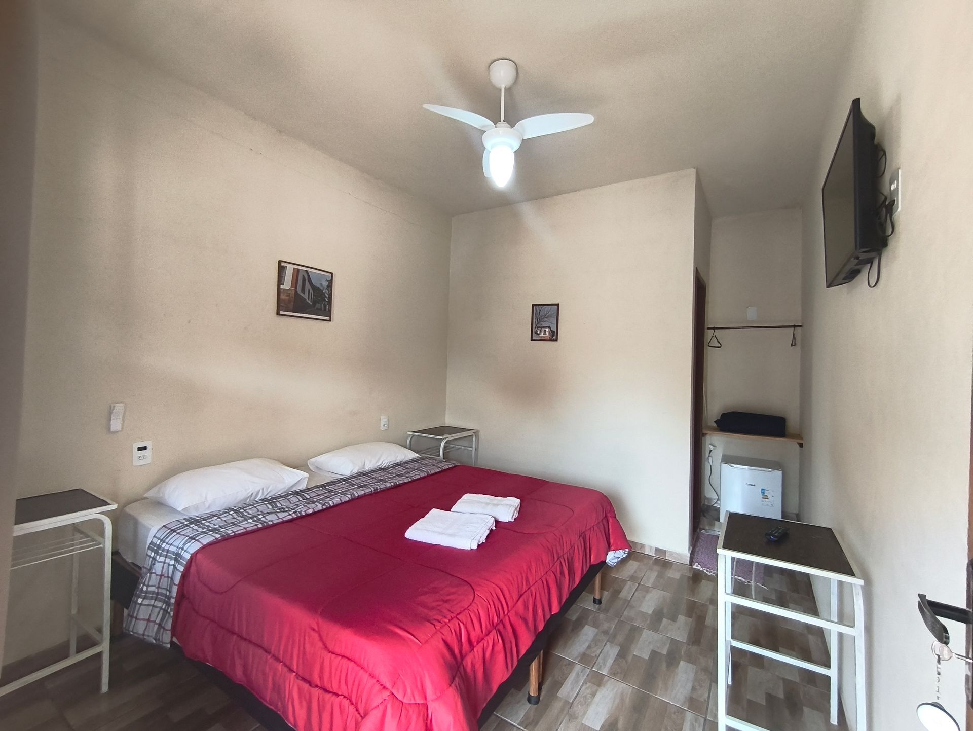 Quarto com colcha vermelha, duas mesas de cabeceira, TV e ventilador de teto.