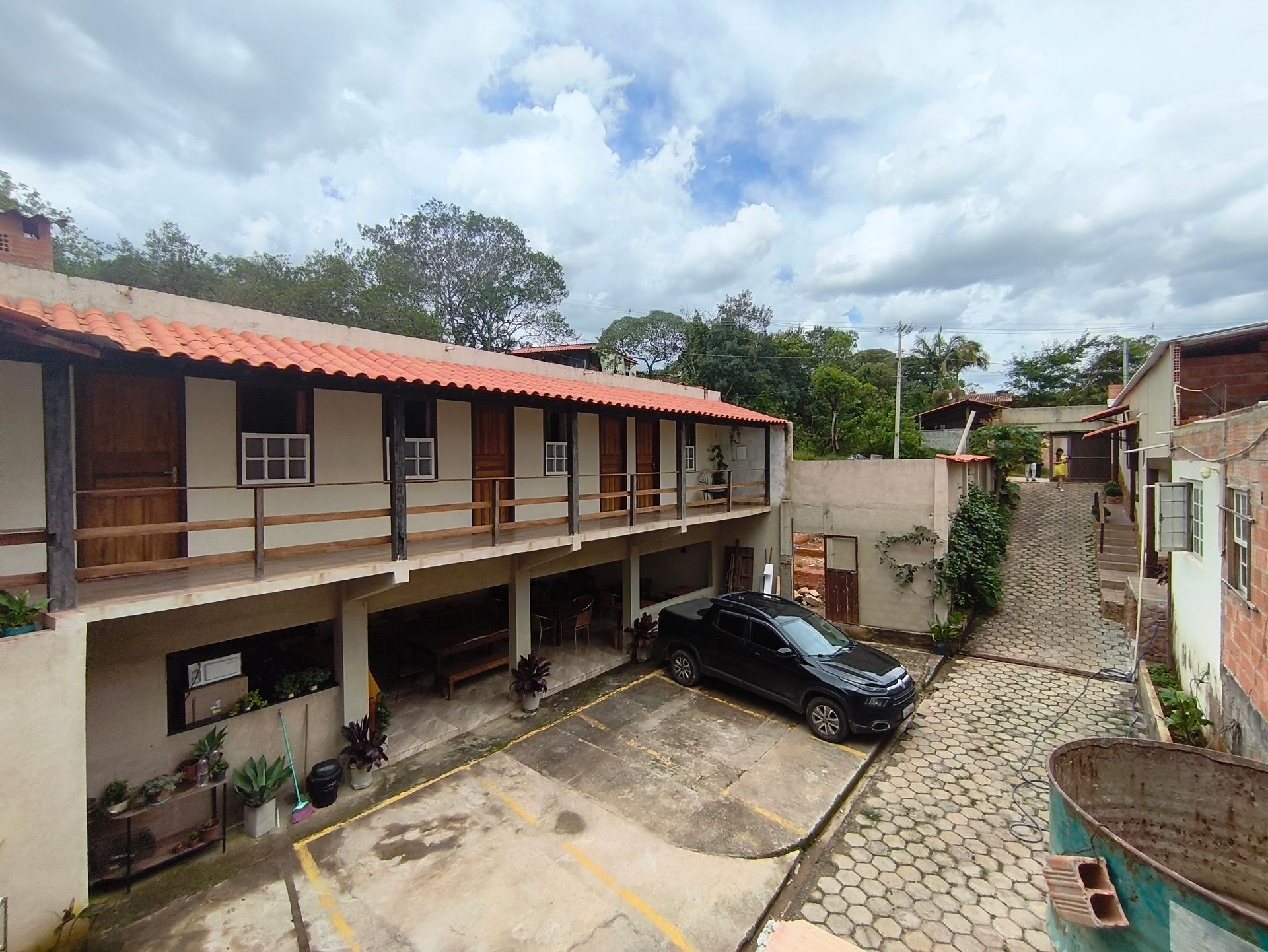 Prédio de dois andares com telhado de telha vermelha, varanda e estacionamento; carro preto estacionado em frente. Céu nublado.