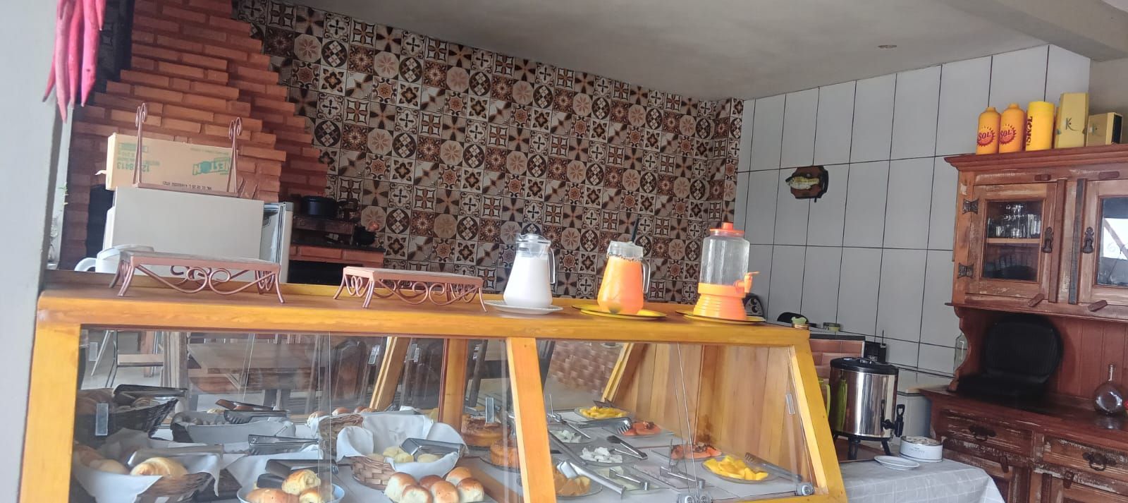 Uma vitrine de padaria repleta de alimentos, bebidas e itens de decoração, encostada em uma parede de tijolos com padrões.