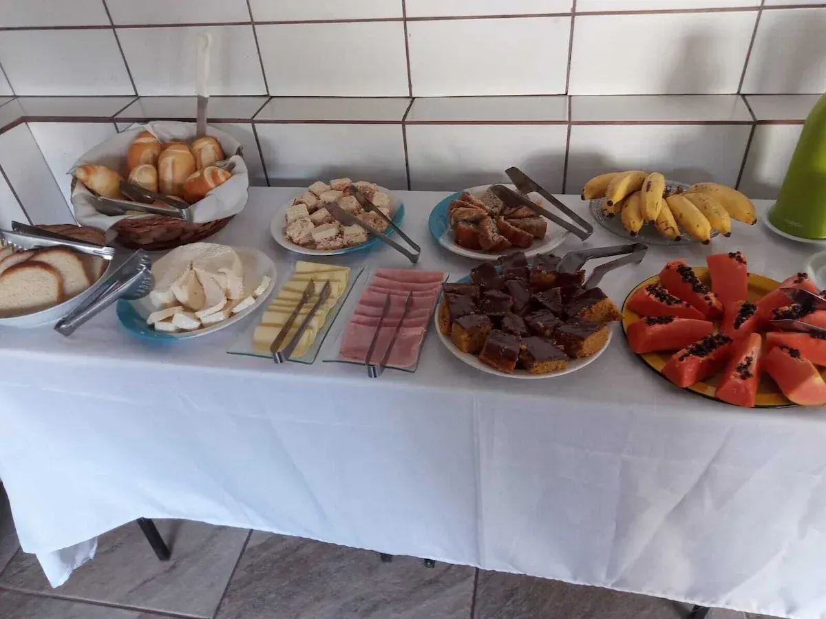 Buffet de café da manhã com diversos doces, frutas e frios, servido sobre uma toalha de mesa branca.
