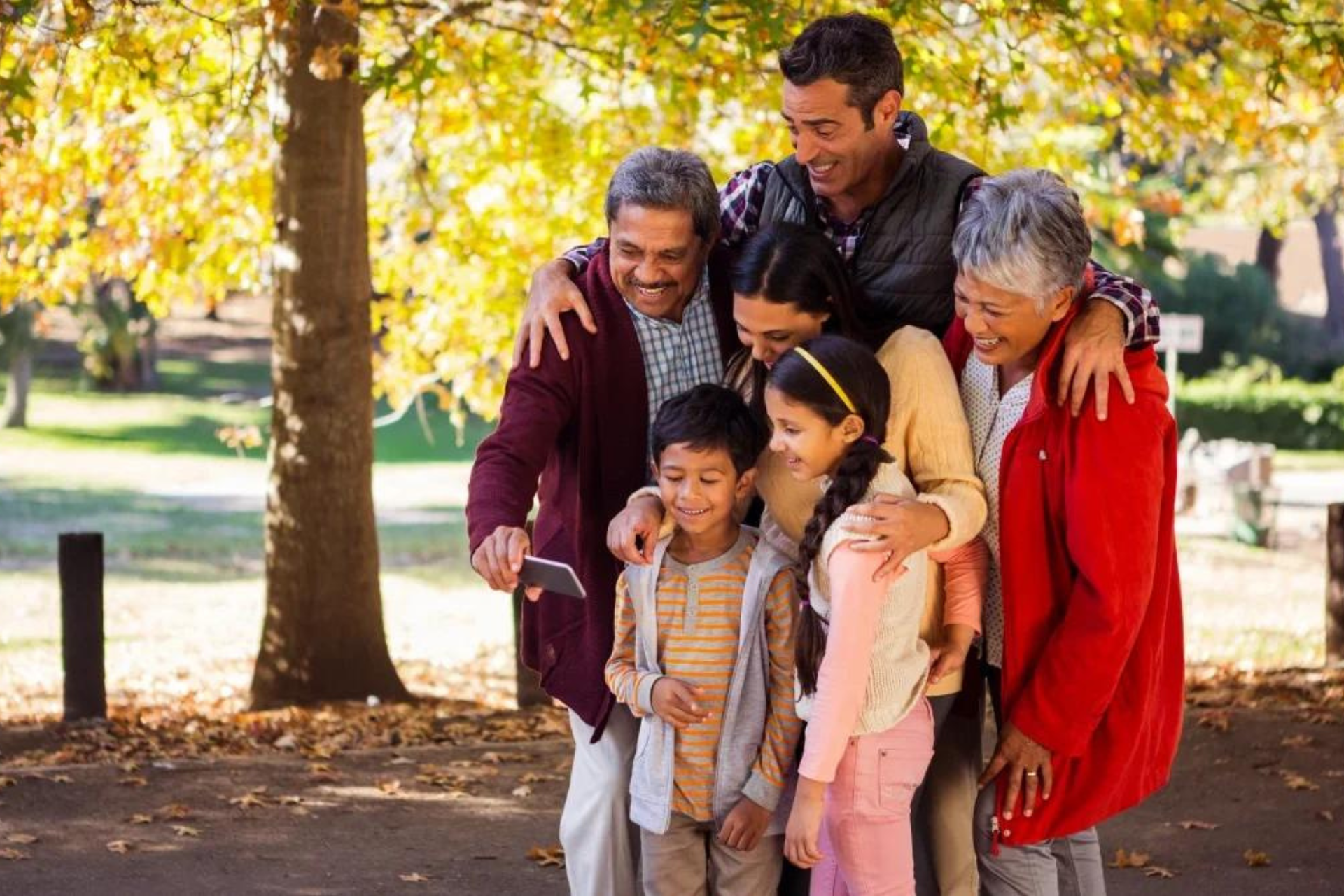 Family of seven smiles together in a park, surrounded by fall foliage; includes children, parents, and grandparents.