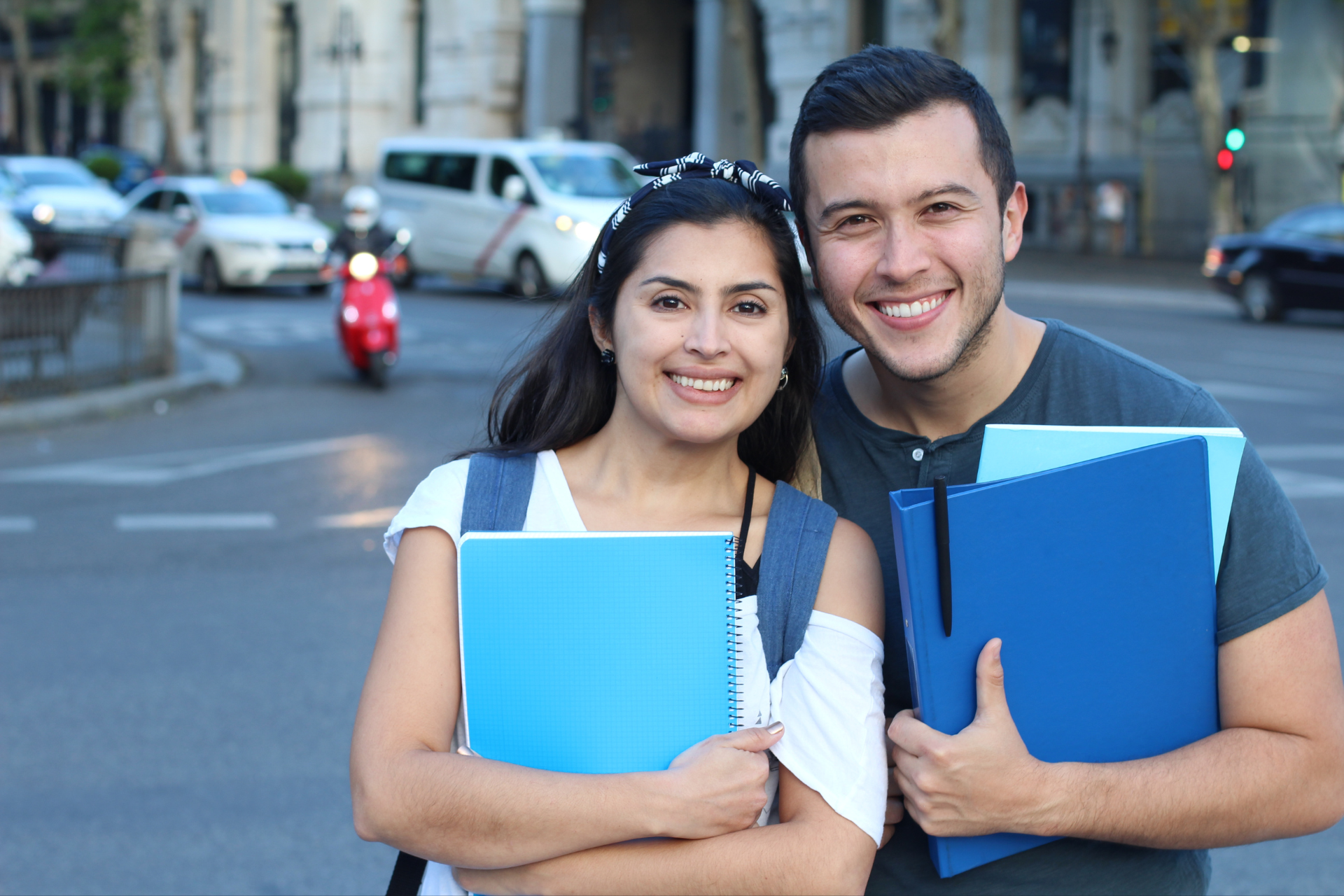 Two smiling students holding blue notebooks on a city street with traffic and buildings in the background.