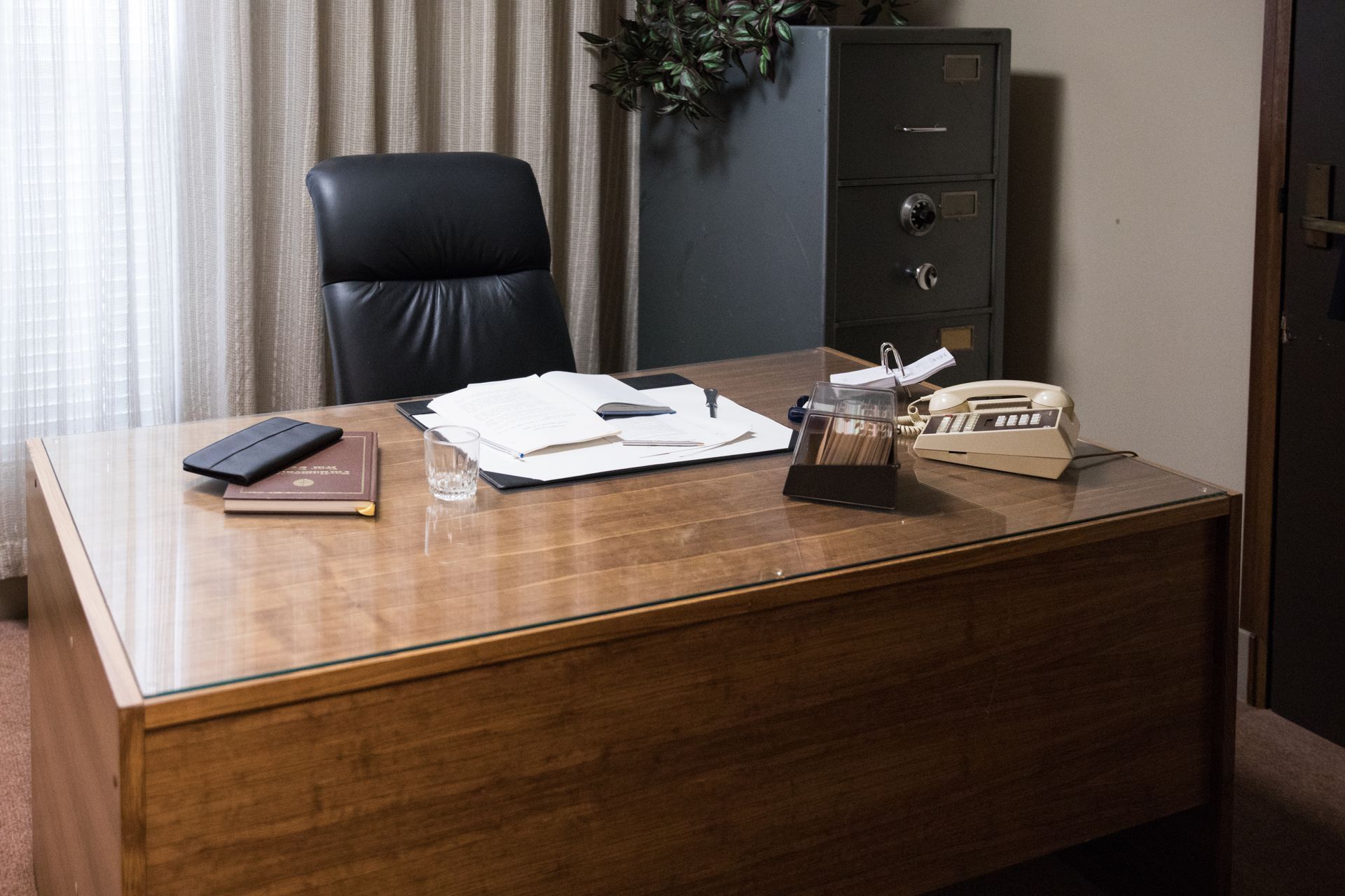 A vintage office with a wooden desk, leather chair, and a safe. Books and a telephone sit on the desk.