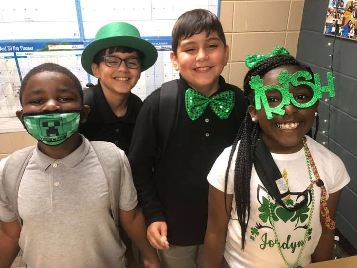 Four students smiling, wearing St. Patrick's Day accessories in a classroom setting.