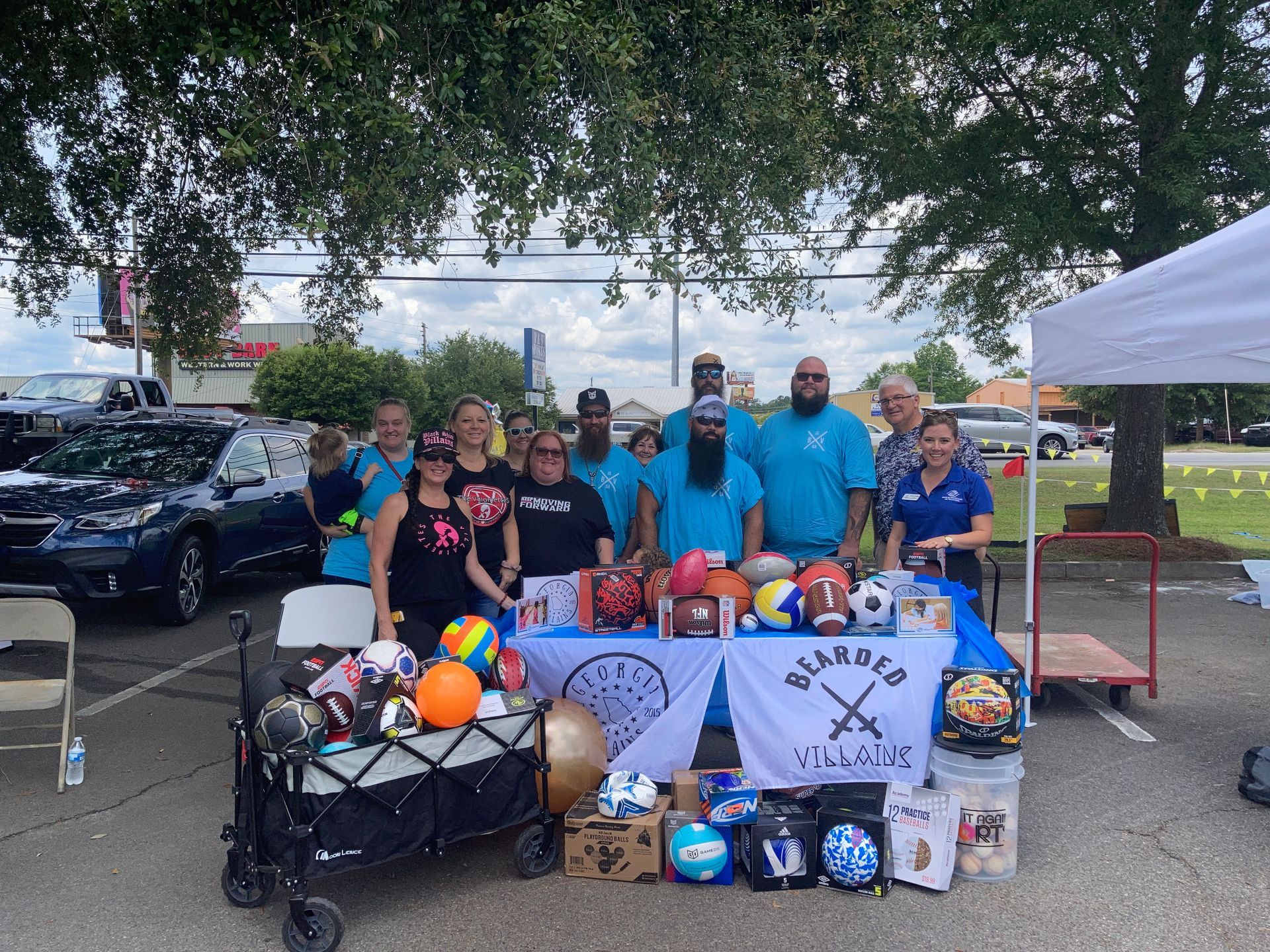 A group of people at a table with sports balls and gear, outdoors. Some wear blue shirts under a tree.