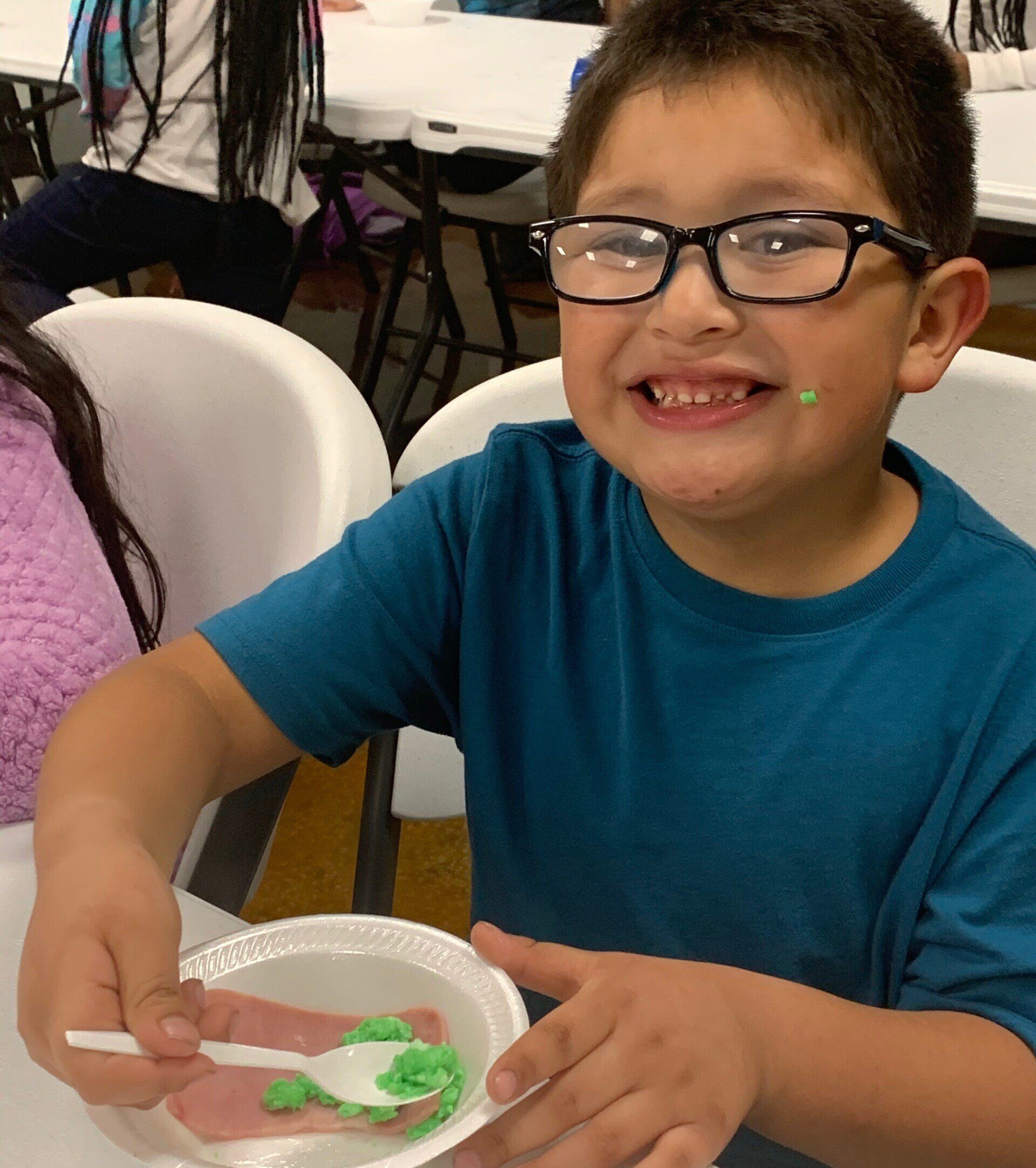 A smiling boy with glasses, a blue shirt, and green frosting on his face, eating from a white plate.