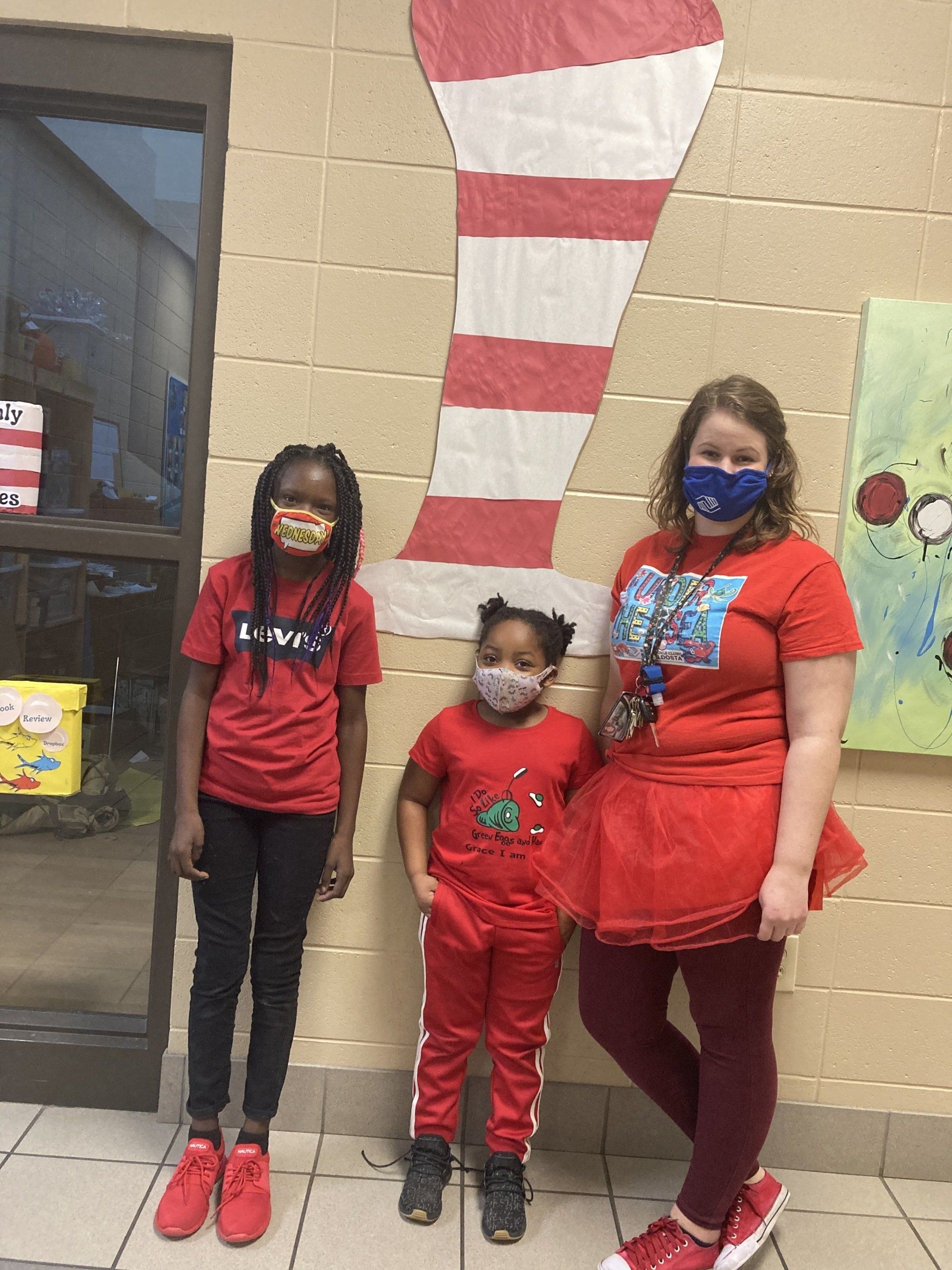 Three people in red outfits and masks stand in front of a red and white striped hat prop.