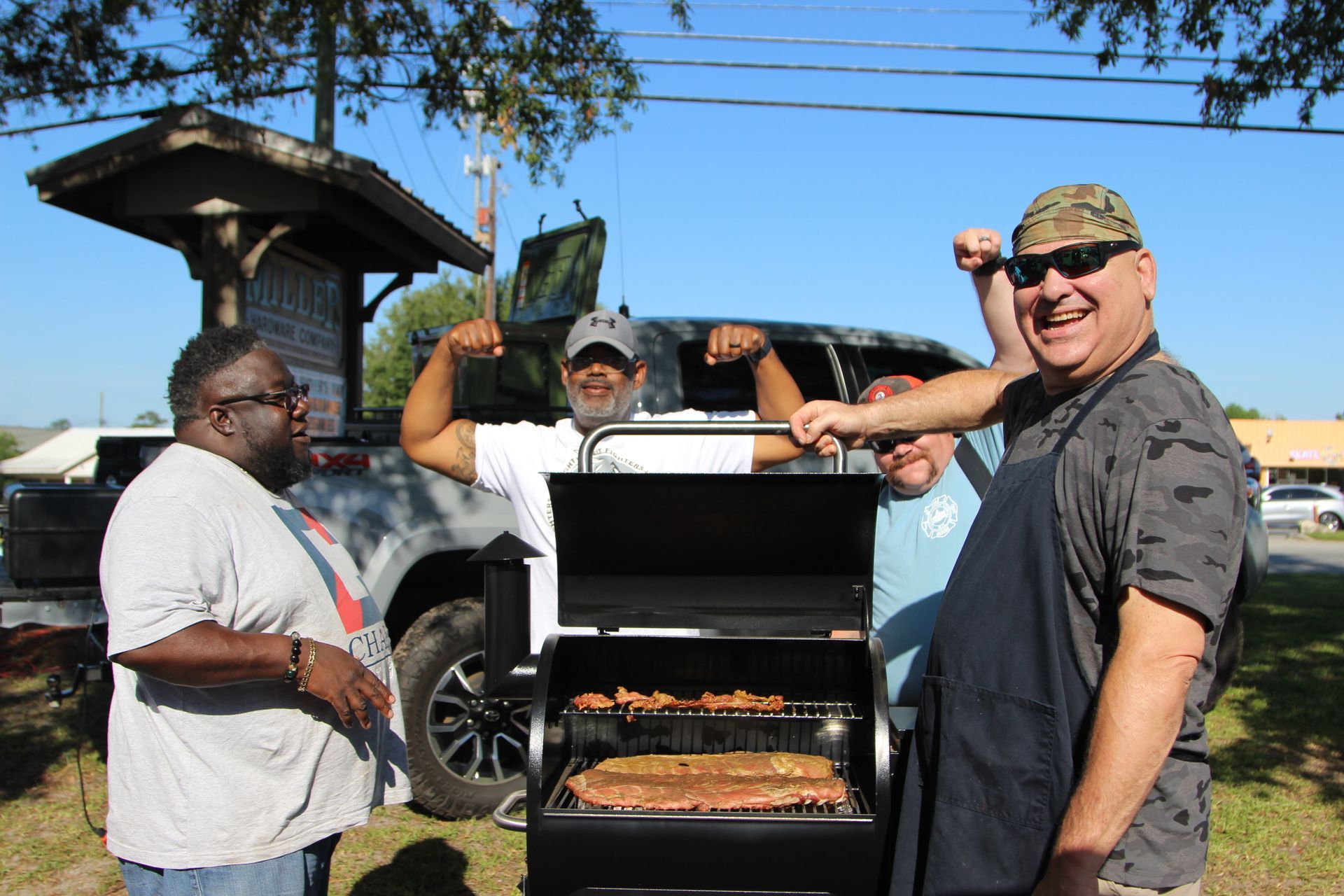 Four men around a smoker grill outdoors. One flexes, smiling; others watch, smiling, on a sunny day.