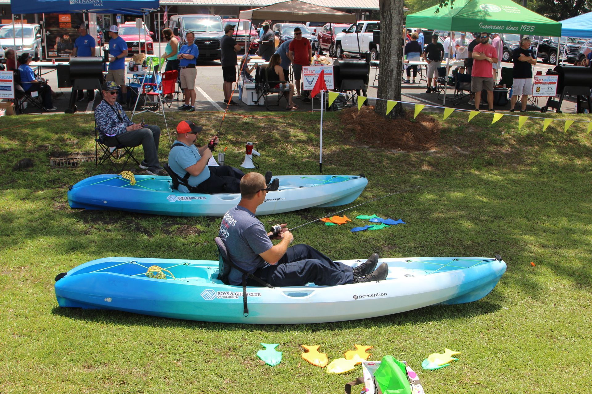 Two men in kayaks on grass, at an outdoor event. People watch in the background.