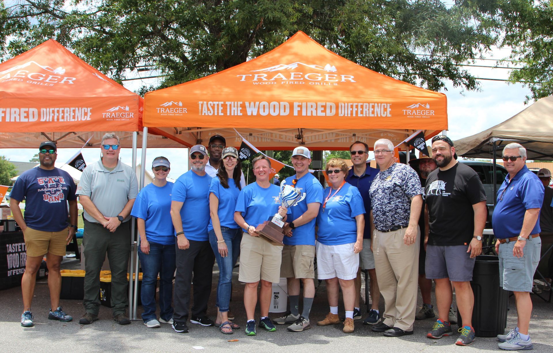 A group of people holding a trophy, standing in front of orange Traeger tents. Outdoors, sunny.