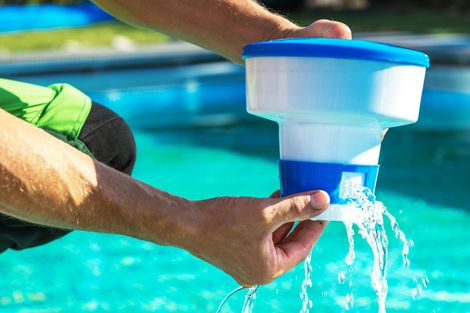 Hands holding a white and blue plastic pool chlorine floater, with water draining from its base into a blue swimming pool.